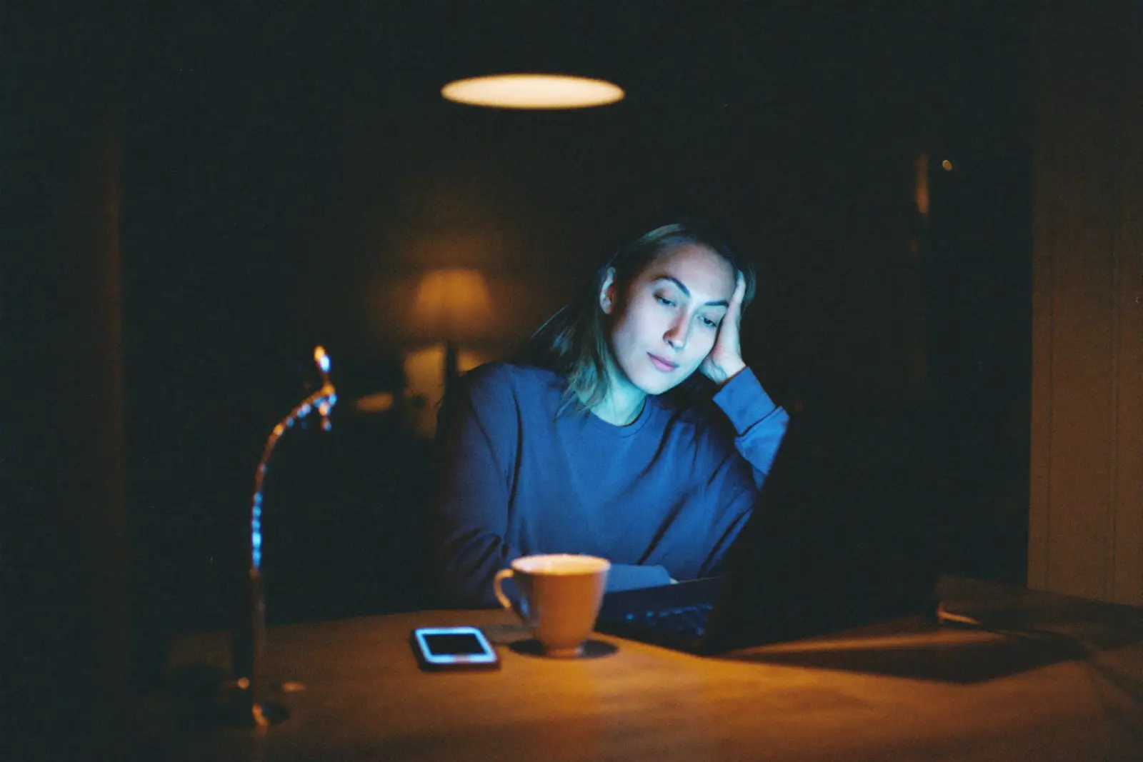A woman works at a kitchen island late at night, her face lit by a laptop screen, phone on the counter beside her