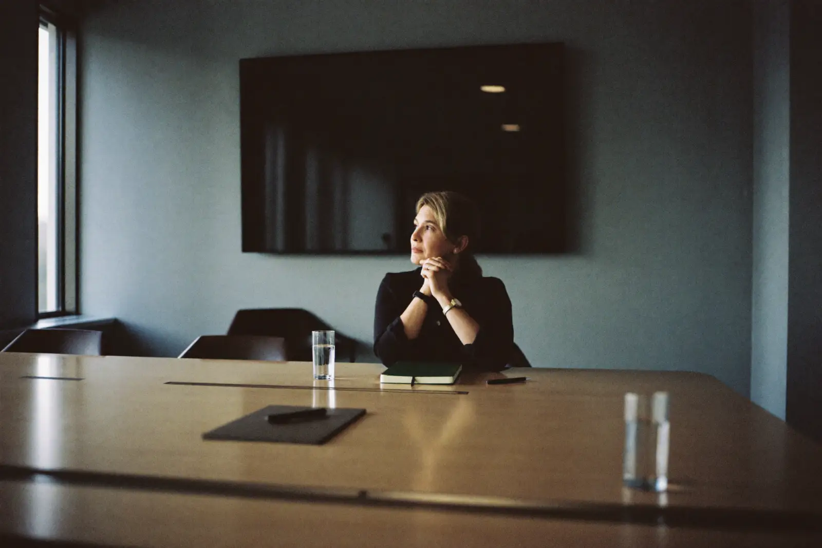 A consultant sits alone in a boardroom, chin on her hands, waiting at a table where the seats opposite her are empty