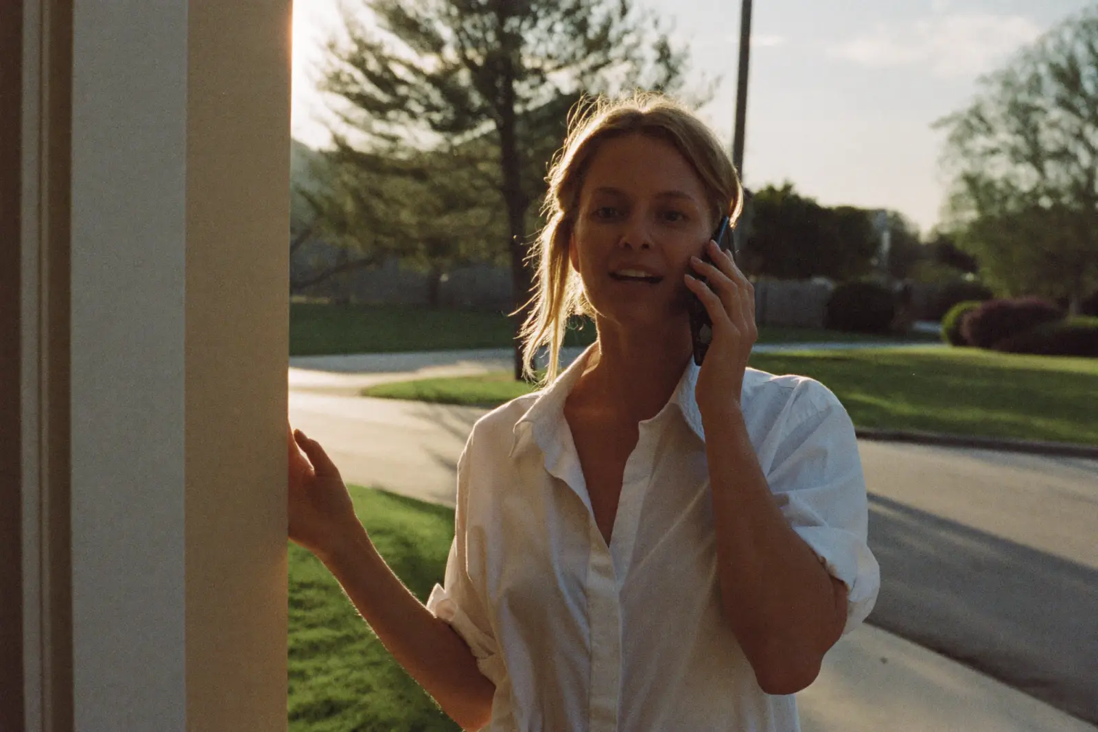 A woman standing outside her front door at golden hour, talking on her phone mid-conversation
