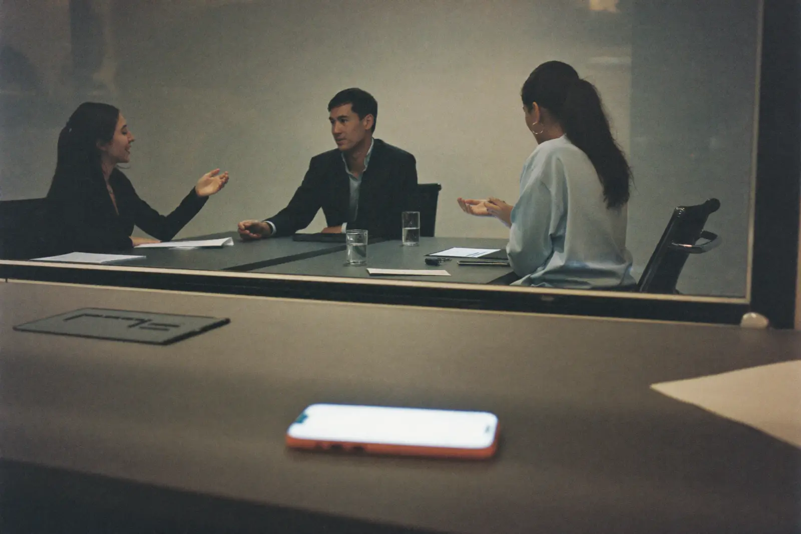 A marketing team works in a glass meeting room while a phone glows unattended on a desk in the foreground
