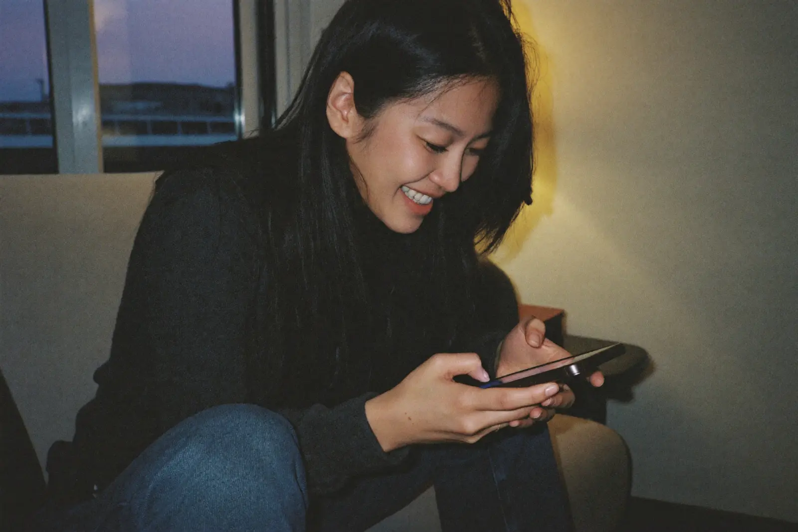 A woman smiling at her phone as she reads a text message in a living room at dusk