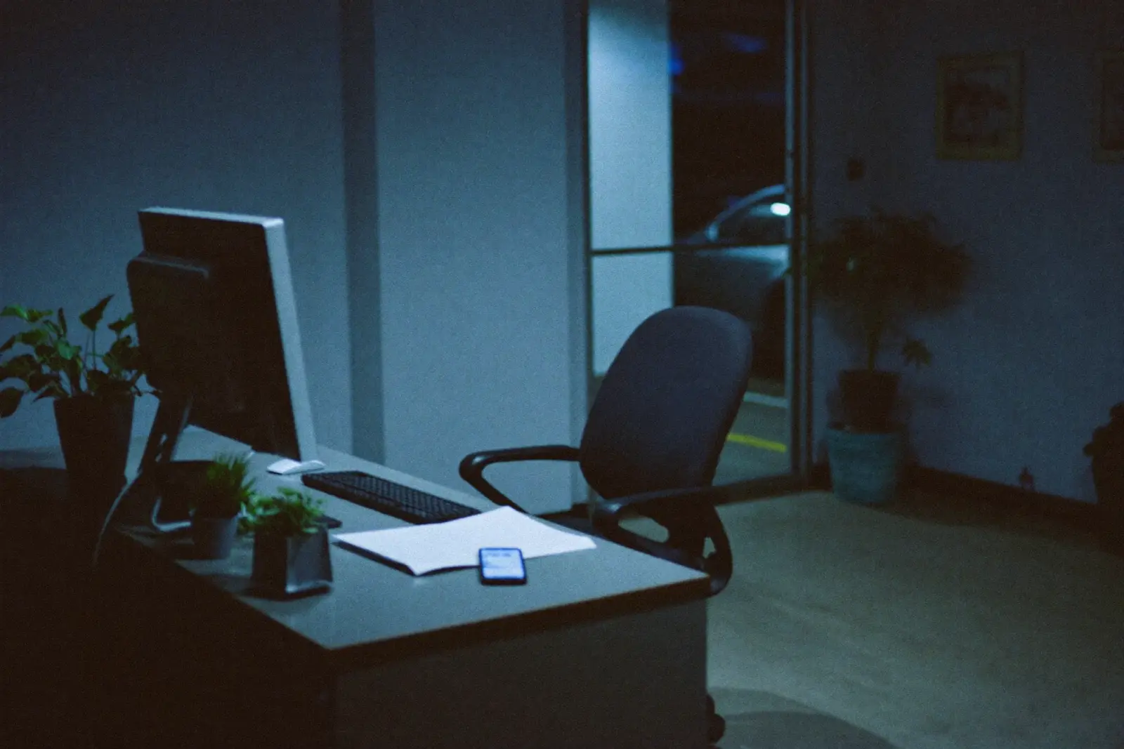 A closed small-business office at night, monitor off and chair empty, with a single smartphone glowing blue on the reception desk